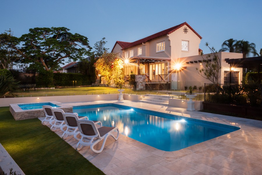 A well-lit house exterior with a yard, swimming pool, and lawn chairs.