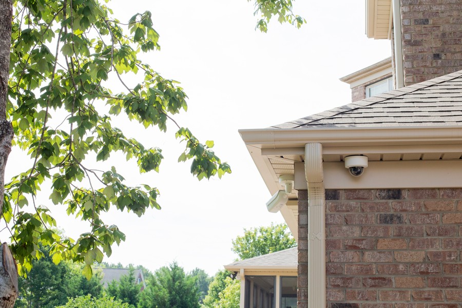 A security camera is shown mounted under the eave of a brick home.