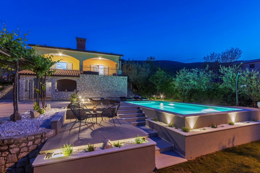 Luxury backyard featuring low-voltage landscape lighting around a modern pool, patio seating area, and stone-accented home at dusk.